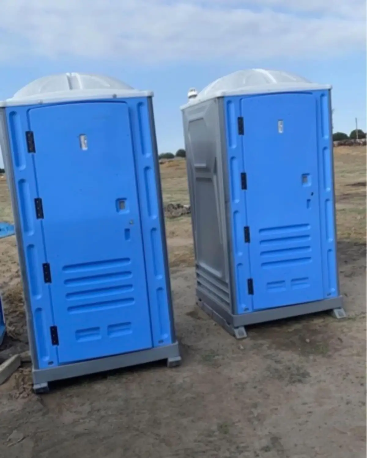 Two blue standard portable toilets set on a dirt construction lot under a clear Texas sky